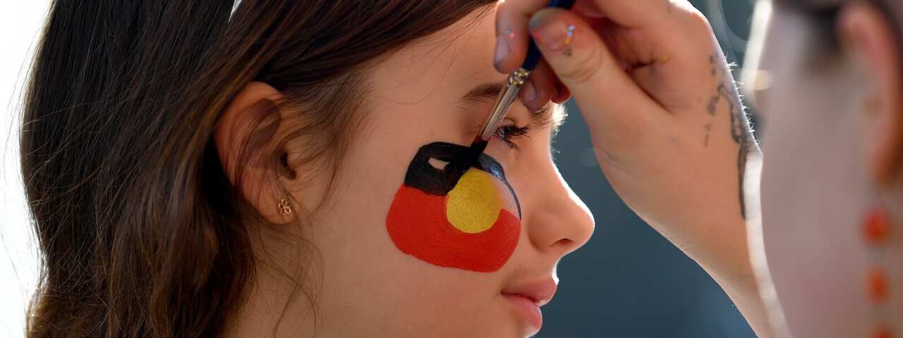 A girl having her face painted with the Australian Aboriginal flag.