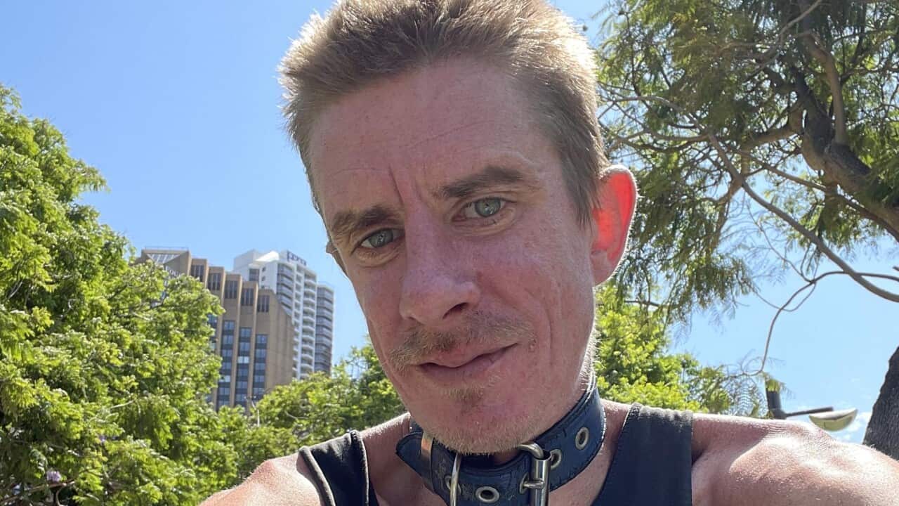 A man wearing a black singlet and a leather collar on his neck takes a selfie outside on a sunny day.