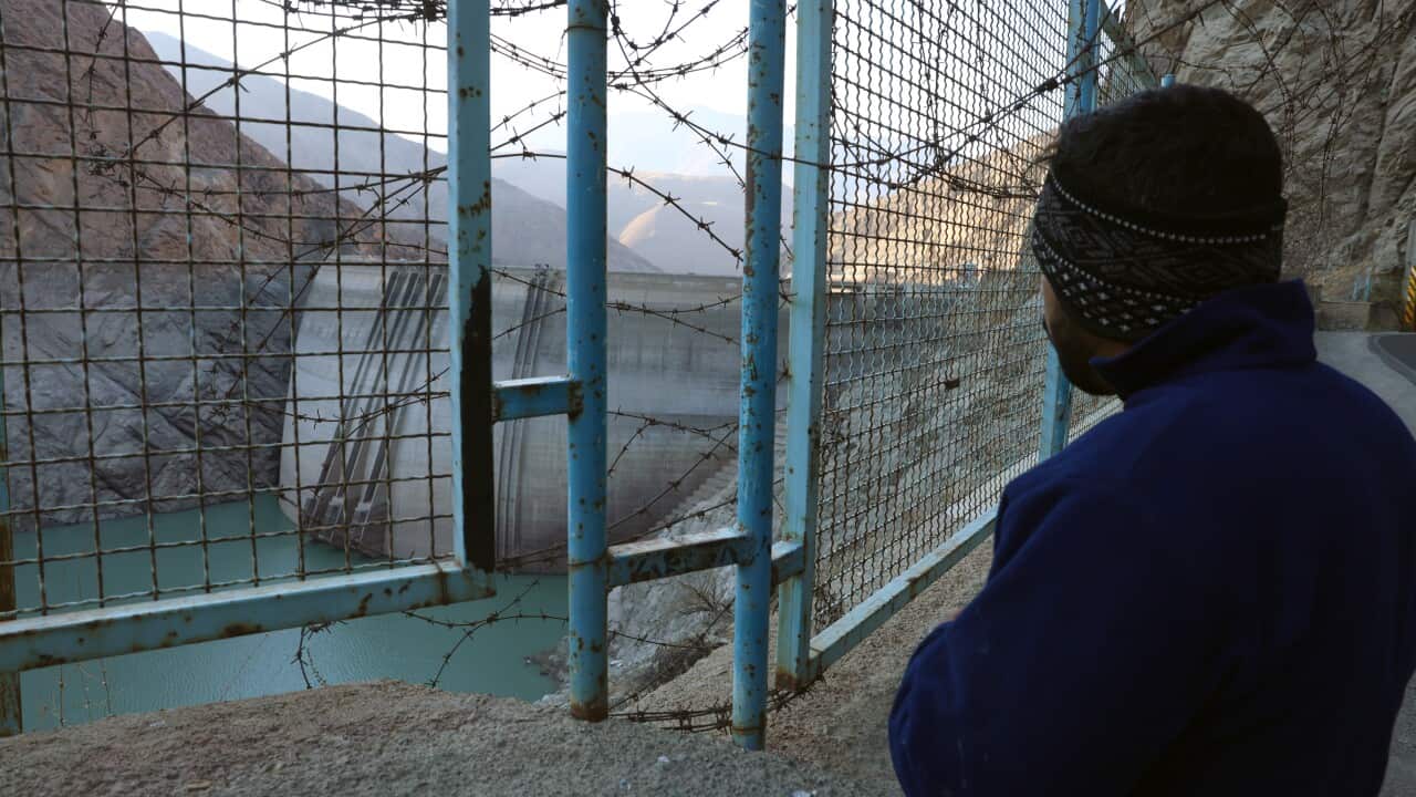 Man looking through a barbed-wire fence at a dam and low reservoir.”