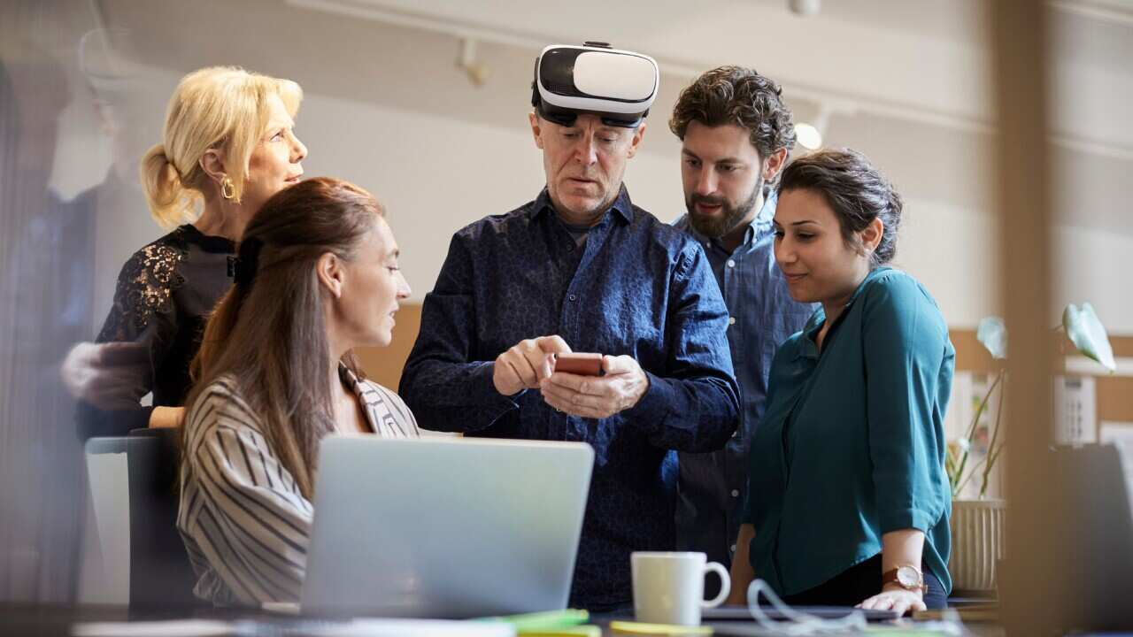 Mature businessman with virtual reality simulator using smart phone while standing amidst colleagues