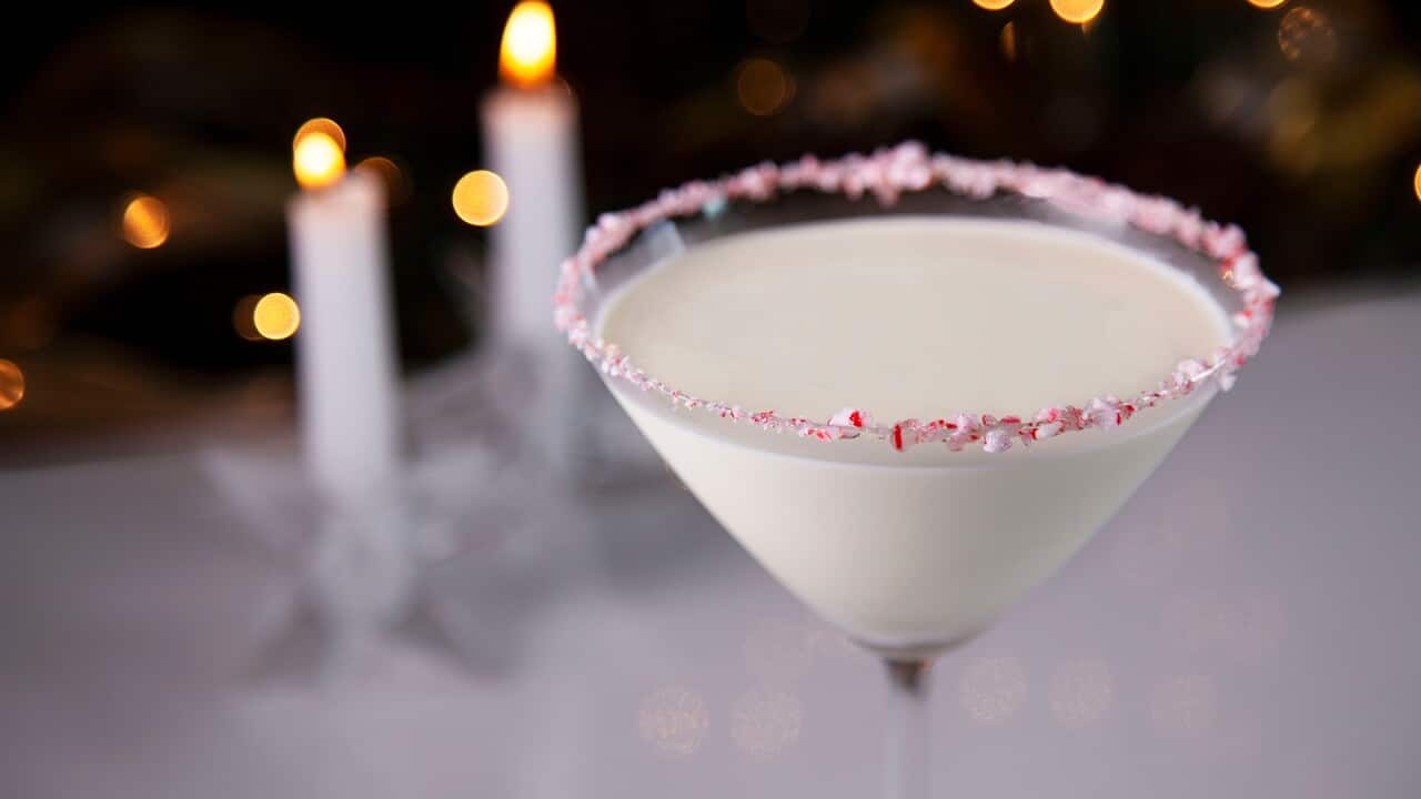 A martini glass rimmed with crushed candy cane holds a white cocktail. Two lit candles can be seen behind the glass, out of focus.
