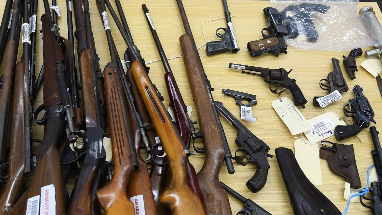 A collection of various firearms, including rifles and handguns, laid out on a table. Some of the long guns have tags on them.
