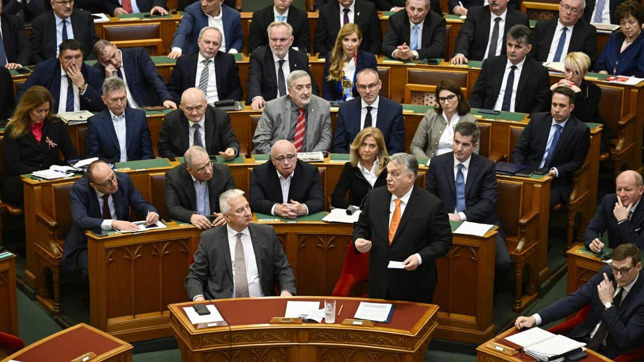 Room full of wooden desks, each placed on tiered levels. People in business wear and suits are sitting at the desks, with one man at the front with a black suit and orange tie standing and delivering an address.