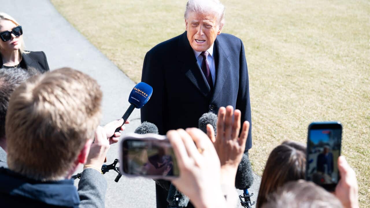 An older man, wearing a dark overcoat and red tie, speaks to reporters outdoors while several journalists hold microphones and smartphones toward him.