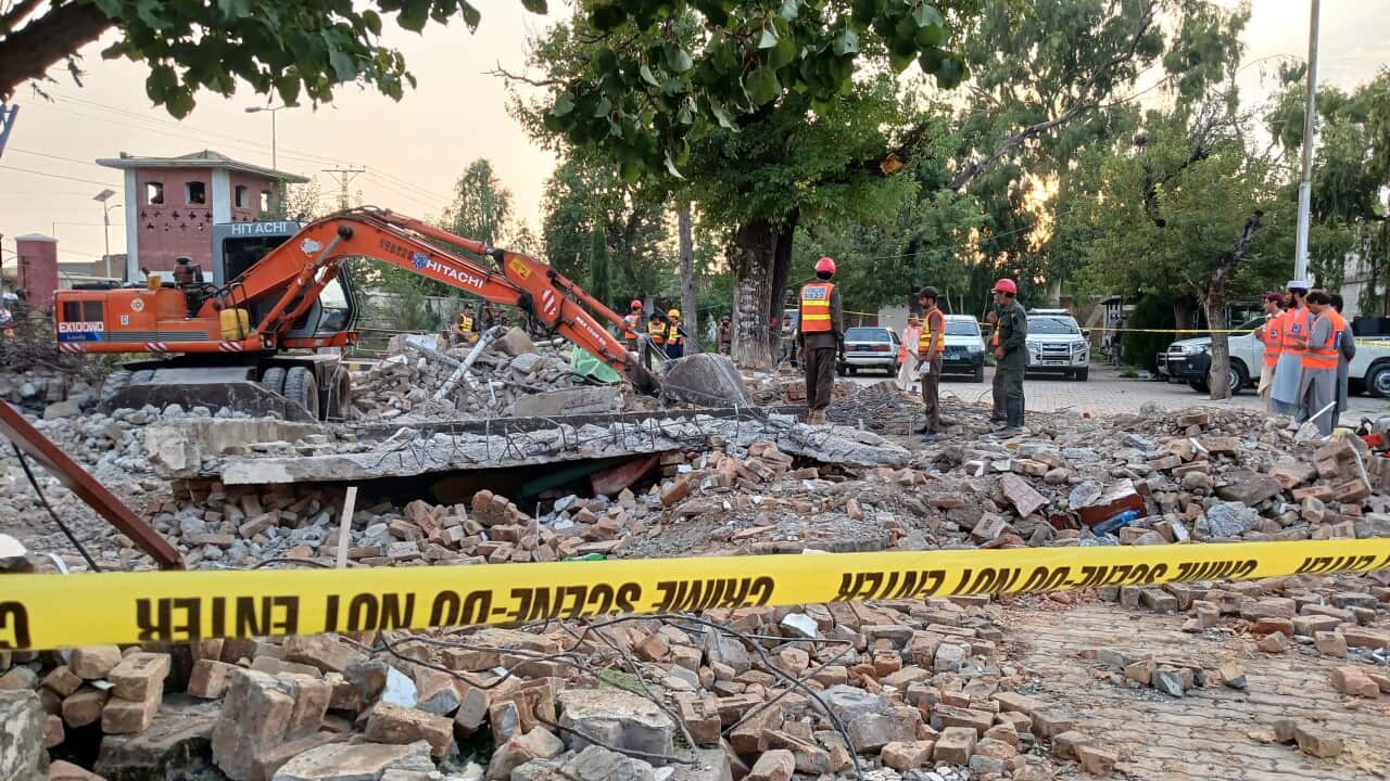 Rescuers search the rubble of a destroyed building, marked off by police tape, with a crane in the background.