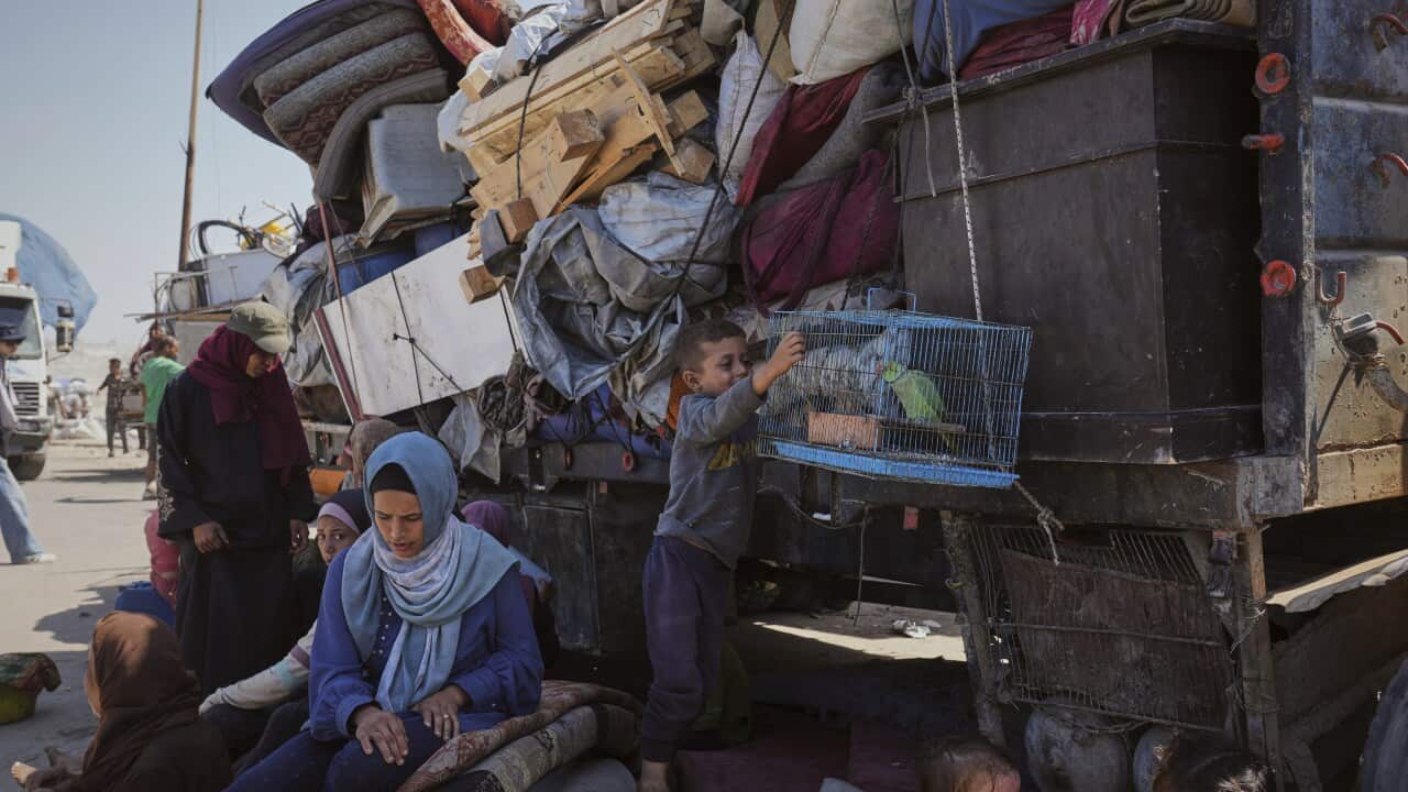 A woman in a blue headscarf sitting on a carpet, and a boy in blue holding a cage with a bird in it. A truck is piled up with luggage behind them.