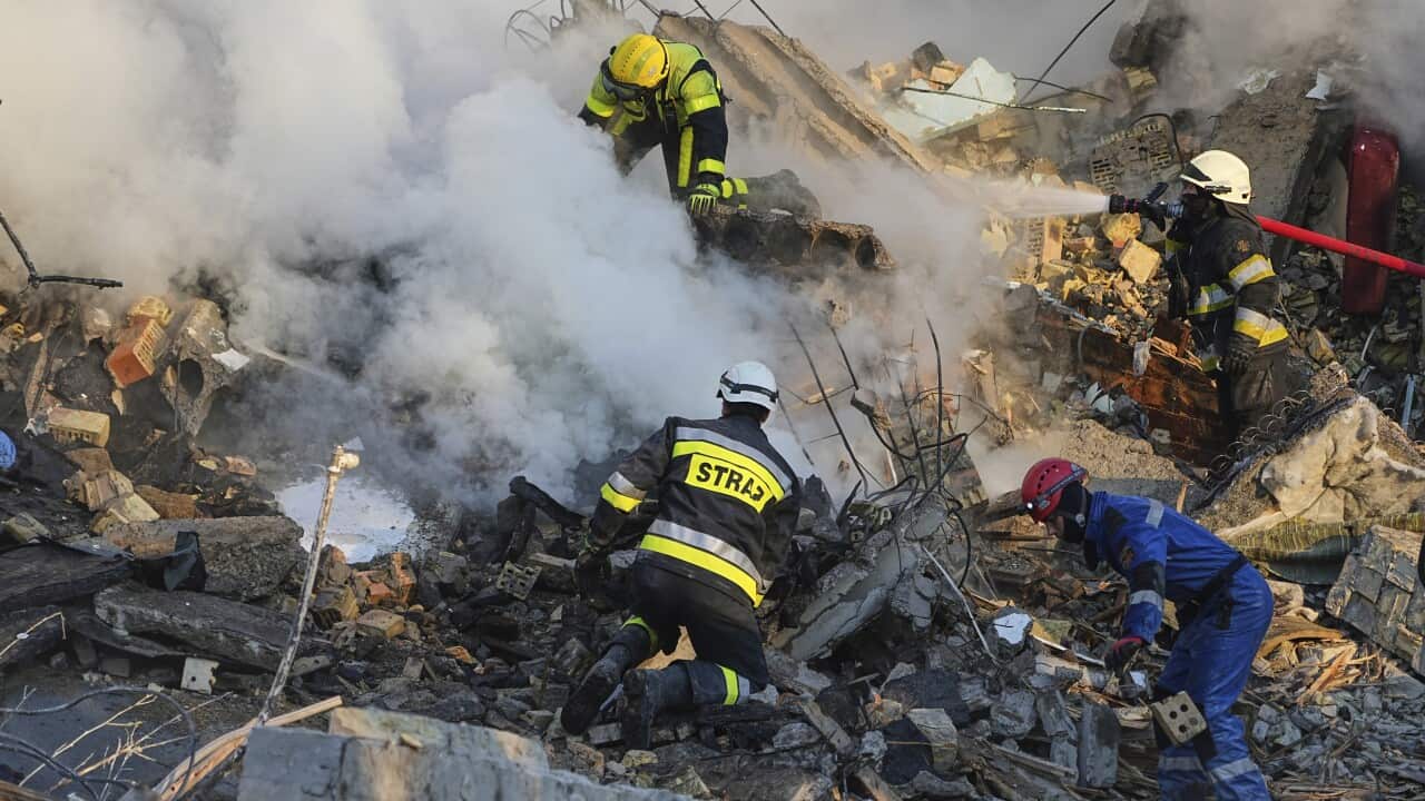 A wide-angle, eye-level shot shows four firefighters in full gear working on a pile of rubble and debris. One is spraying a hose into a cloud of white smoke.