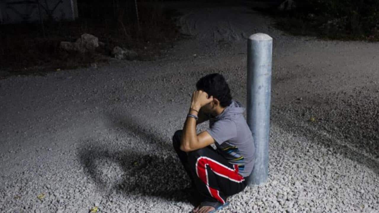 A man squats down leaning his back against a pole.