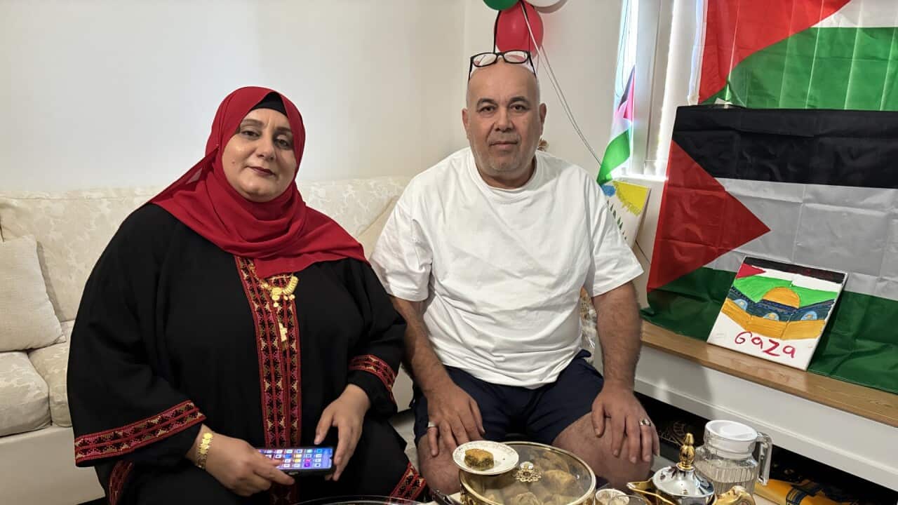 A couple is seated in a living room, with a woman in a black and red embroidered dress and hijab holding a phone, next to a man in a white t-shirt and dark shorts. They are positioned behind a large platter of baklava and other sweets, with a Palestinian flag visible in the background.