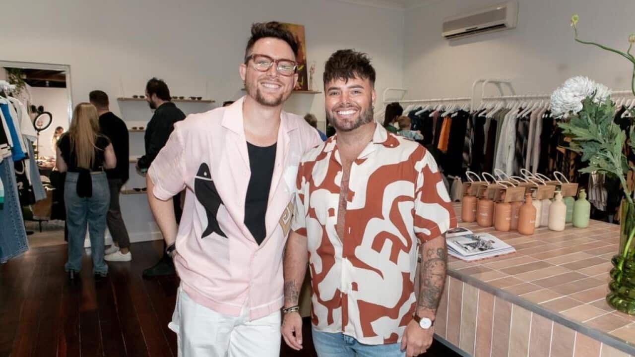 Two men smile at the camera in a clothing shop. The man on the left is wearing a pink shirt and glasses and the man on the right is wearing an orange and white patterned shirt.