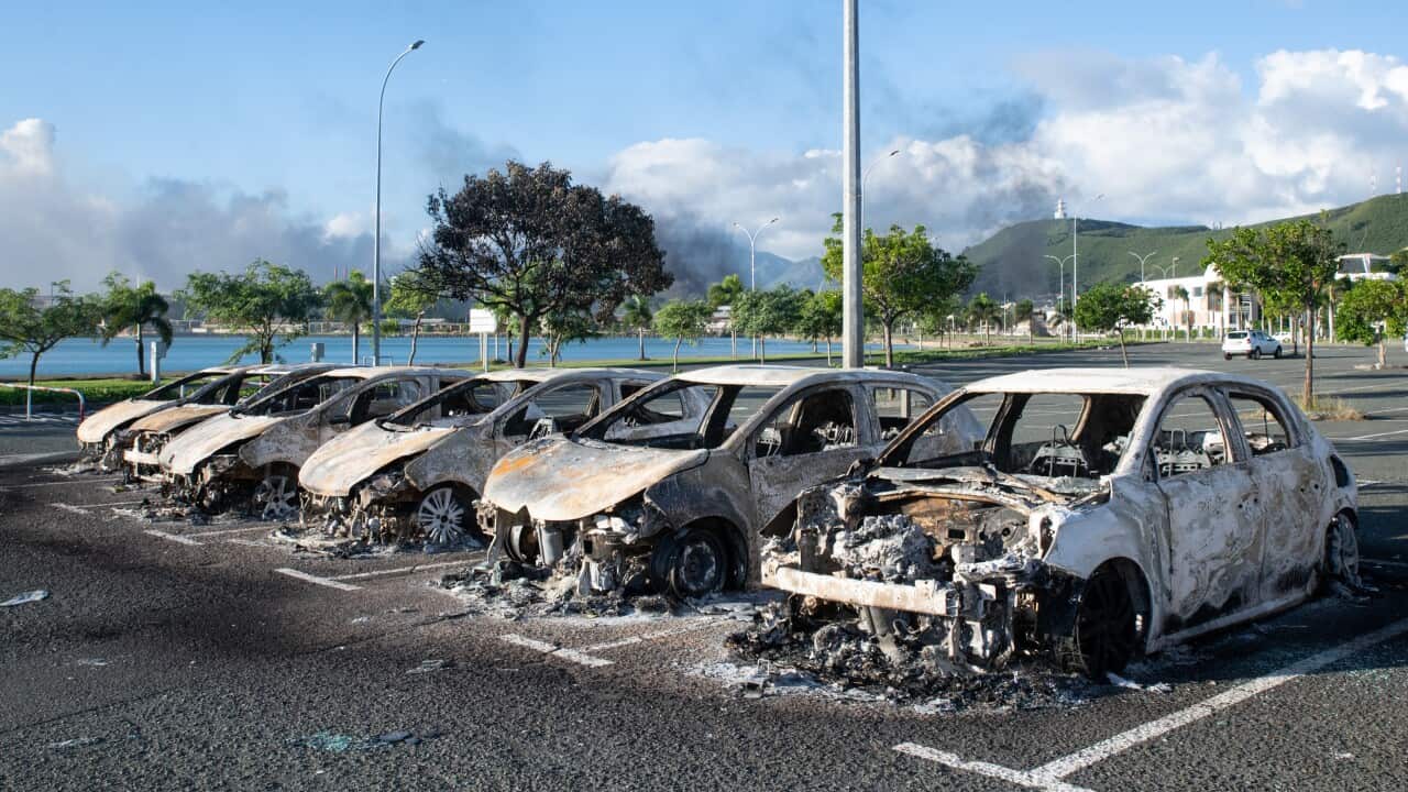 A row of burned-out cars in a parking lot, with a view of the sea and green hill behind them.