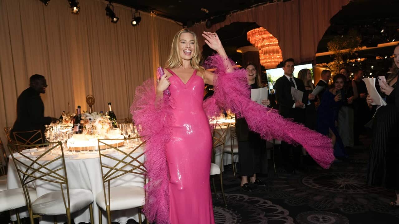 A woman wearing a pink dress smiles and waves while standing next to a table.