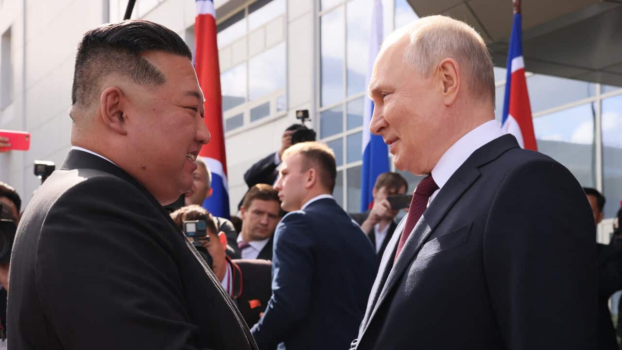 Two men in suits shake hands surrounded by photographers