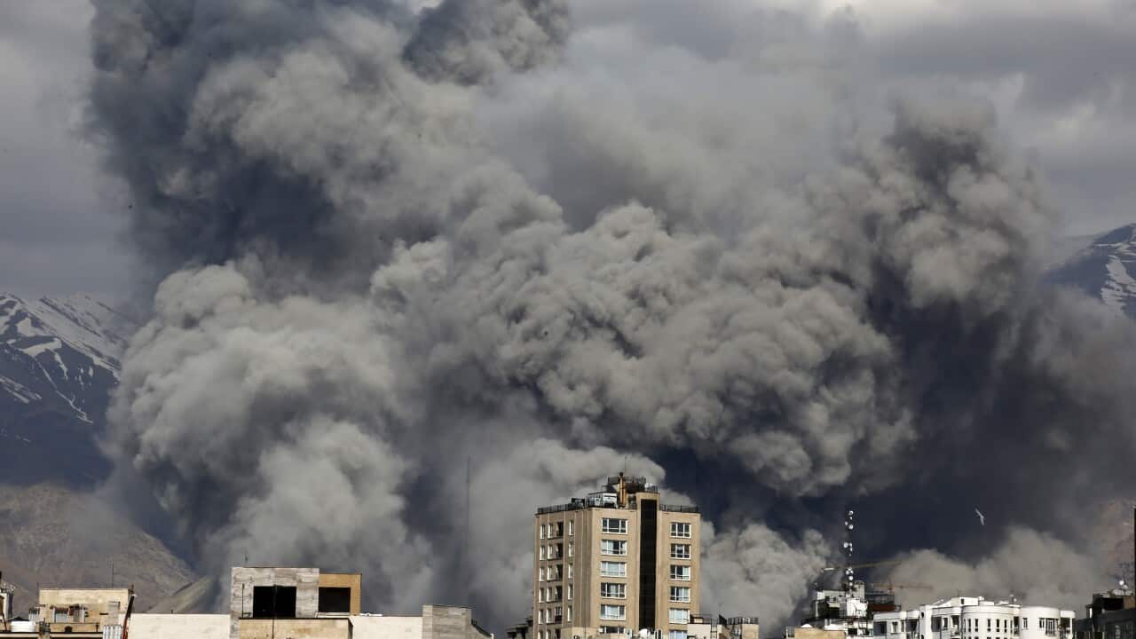 Dense, dark gray smoke billows into the sky above a cityscape, with snow-dusted mountains visible in the background under a heavy overcast.