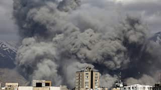 Dense, dark gray smoke billows into the sky above a cityscape, with snow-dusted mountains visible in the background under a heavy overcast.