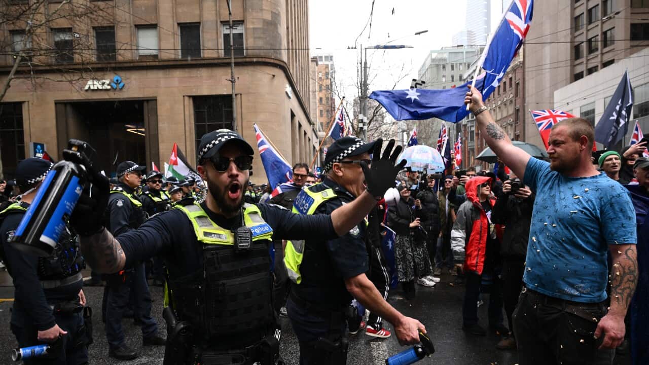 A group of Australian police officers in riot gear are facing a crowd of protestors. One officer on the left is shouting and gesturing with his arms, while another officer is on the ground. A man in the crowd is holding an Australian flag.