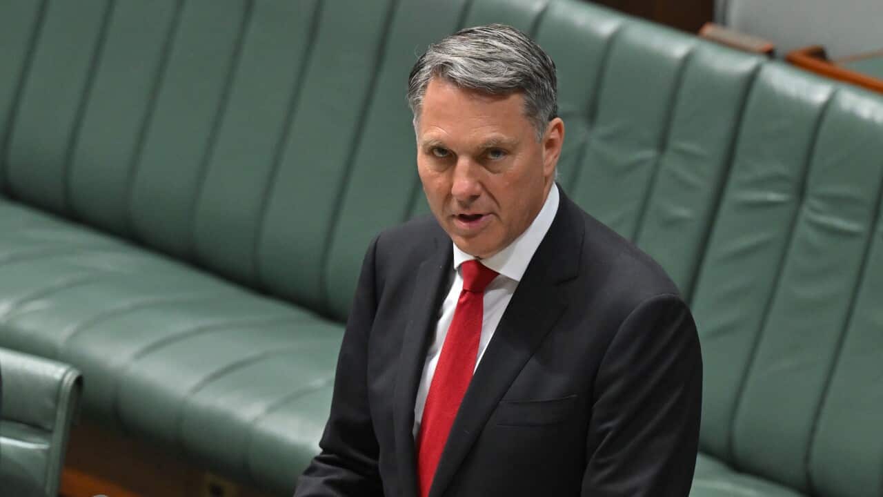 A man stands at a lectern in a suit, reading a speech in Parliament.