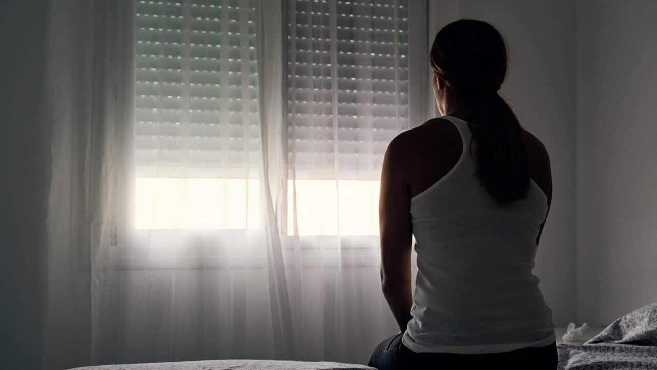 A woman in a white singlet sitting on a bed looking out a window