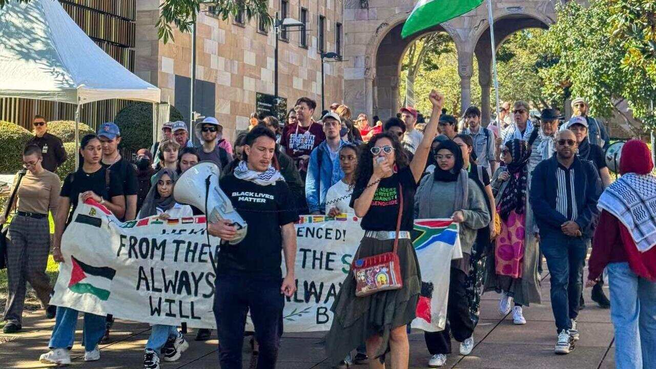 A protest march is taking place on a sunny day, with a crowd of people walking down a path. A woman in the foreground is raising her fist and holding a megaphone, and a large Palestinian flag is visible in the background.