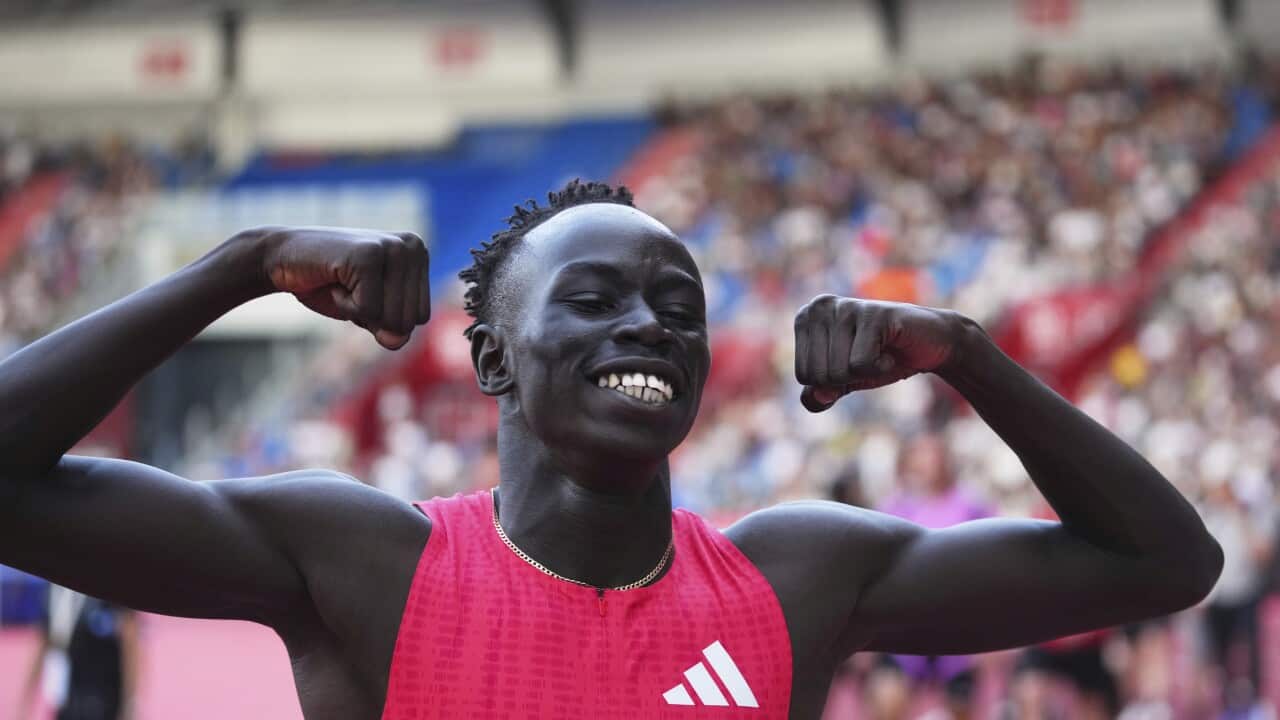 Gout Gout, in a red jersey with a white logo, smiles as he raises his arms in a sign of victory inside a stadium.