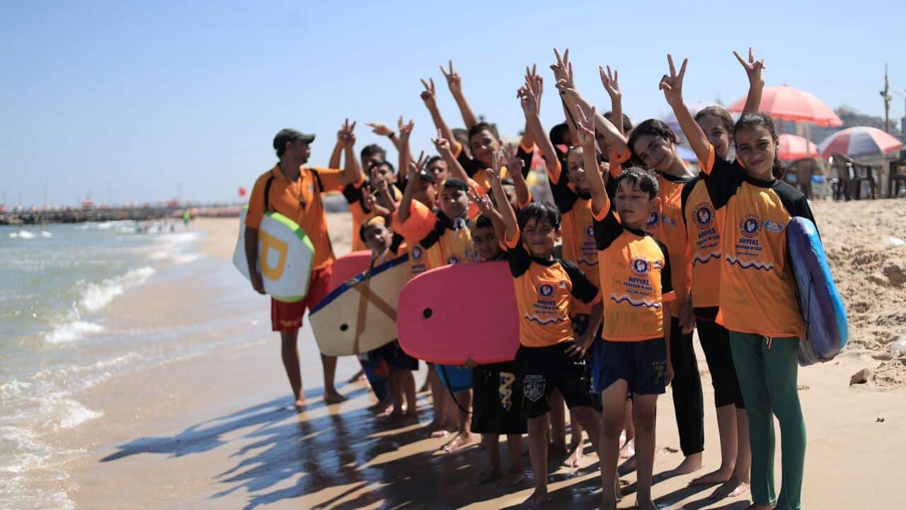 A group of children and one adult instructor stand barefoot on a sandy beach near the ocean, all raising their hands to make V for peace signs. The children are wearing matching orange and black tops and several are holding bodyboards.