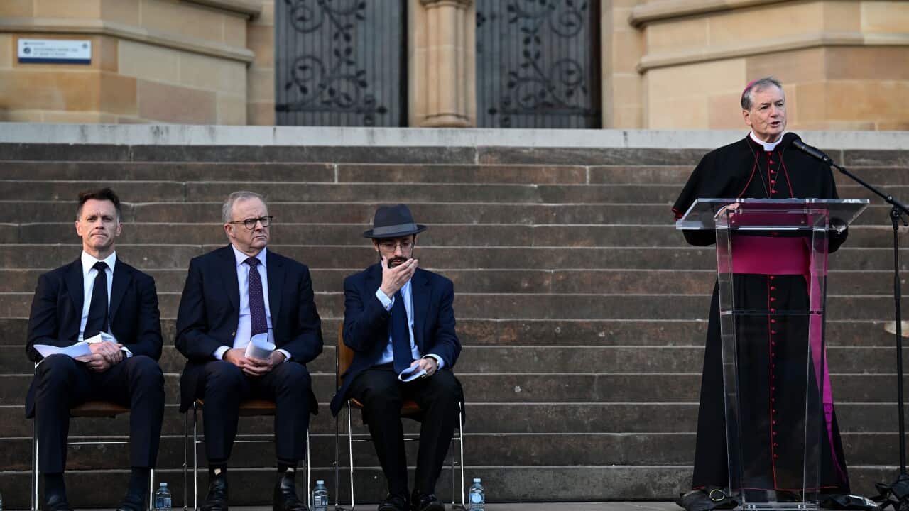 A priest speaks at a podium with three men sitting on his right