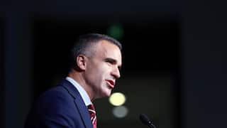 A man in a dark suit with dark hair speaking into a microphone as seen from the side