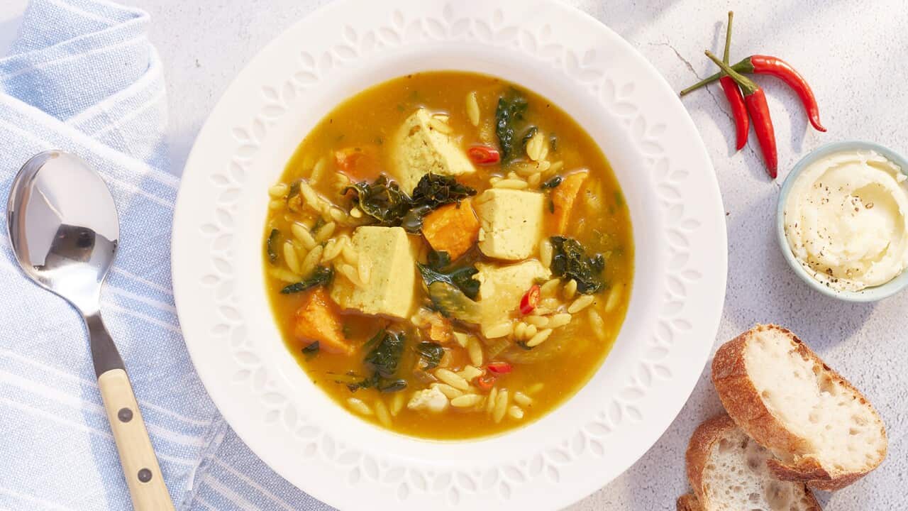 A white bowl with a wide, patterened edge sits on a pale blue striped napkin, on a pale benchtop. Two bowl holds a reddish soup with orzo, vegetables and pieces of tofu.