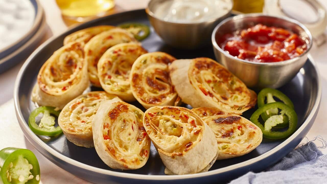 Cooked rolled tortilla rounds with a cheesy filling sit on a tray, along withe bowls of sour cream and red salsa.