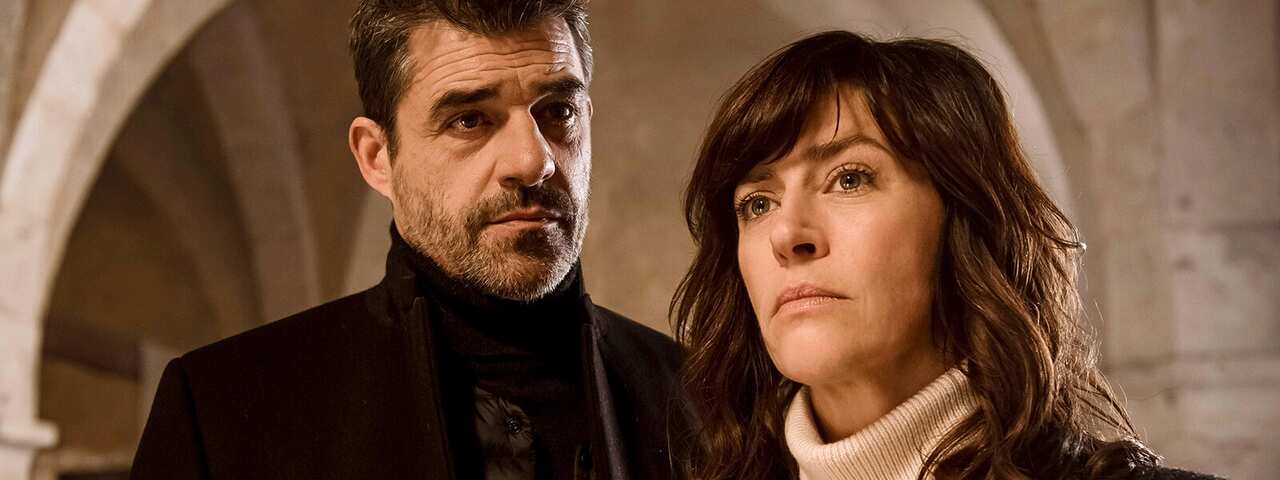 A man and a woman stand in what looks like a church or a historic building, with a stone arch behind them.