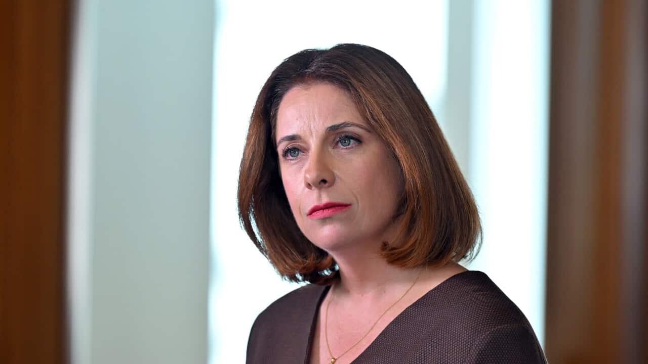 A  headshot captures a fair-skinned woman with medium-length, warm brown hair and intense, light-colored eyes. She wears a brown V-neck top and a simple gold pendant necklace, looking slightly off-camera with a serious expression against a bright, softly blurred background.