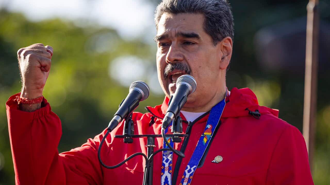 A man in a red jacket and a thick moustache gestures in front of microphones at a podium