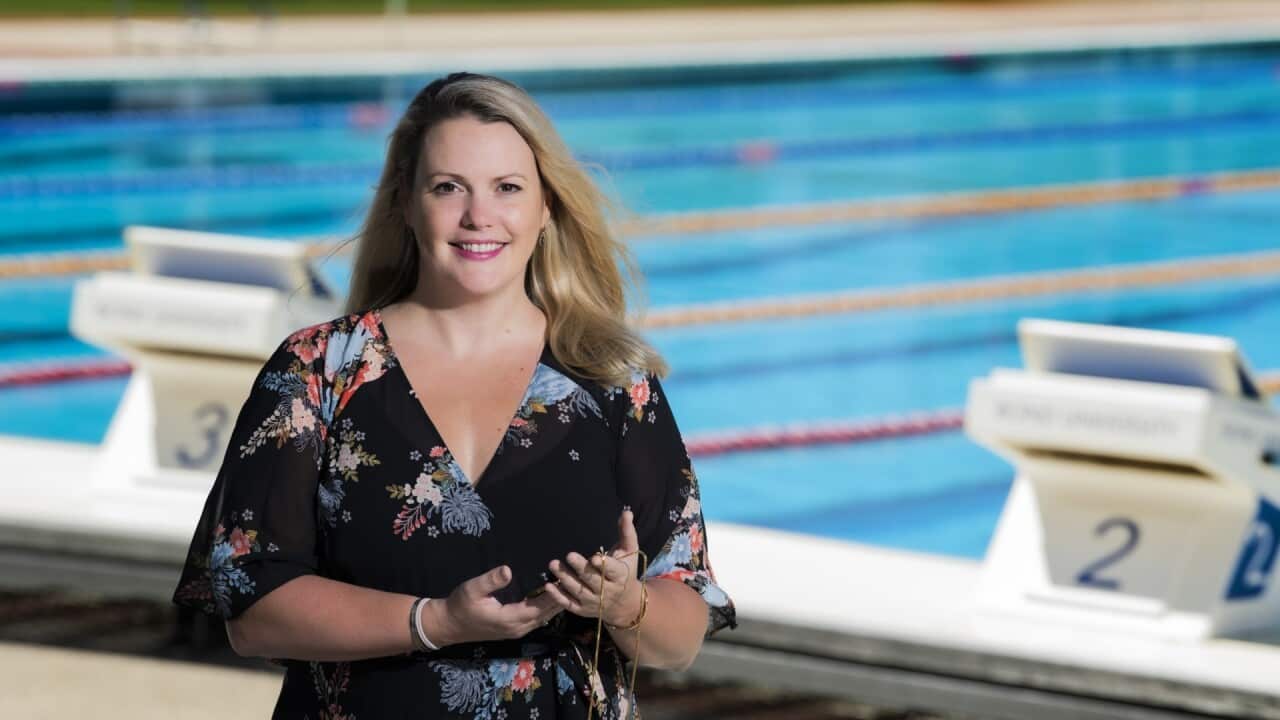 A woman with long blonde hair and a black dress stands outside next to an Olympic-sized pool with a light smile on her face.