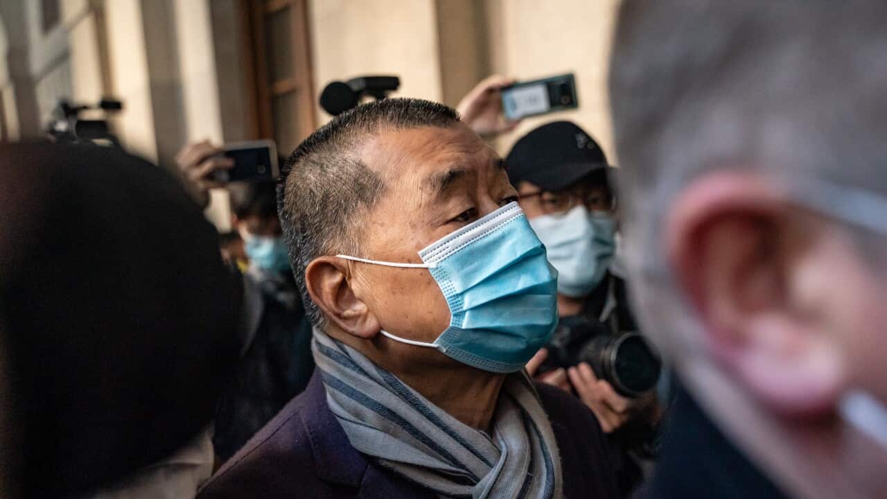 A close-up shot shows Jimmy Lai wearing a light blue surgical mask and a striped scarf as he walks through a crowded area surrounded by people with cameras and smartphones.