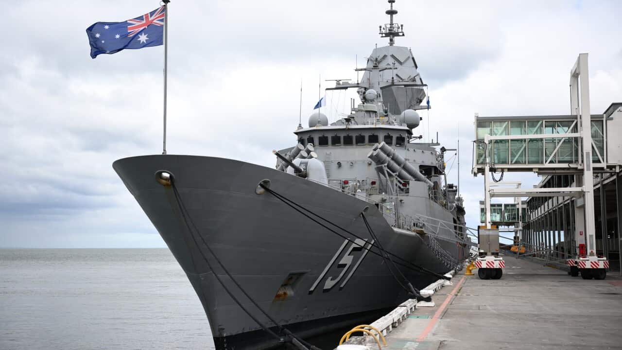 A large grey warship is docked at a pier with an Australian flag flying from its stern.