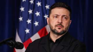 Ukrainian President Volodymyr Zelenskyy stands at a podium in front of a United States flag, appearing sombre and resolute.