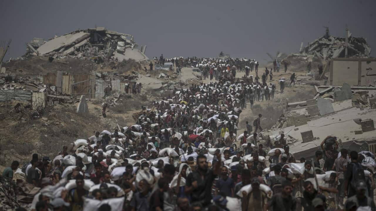 Crowds of people walk carrying bags of flour over their shoulders on a dirt road flanked by destroyed buildings and rubble.