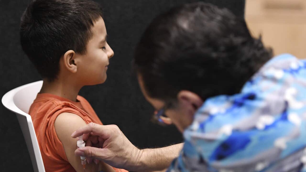 A child in an orange shirt sitting side-on on a white chair closes his eyes as a woman in a blue shirt holds a needle to his bicep. The woman's back is to the camera and she's out of focus.