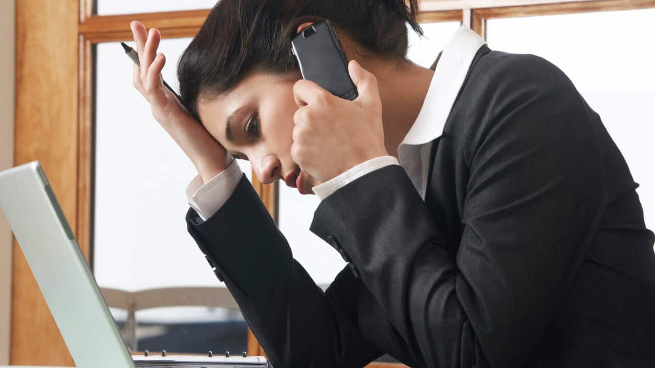 A woman in a black jacket worn over a white shirt, leaning on her hand, looking down at her computer while on the phone.