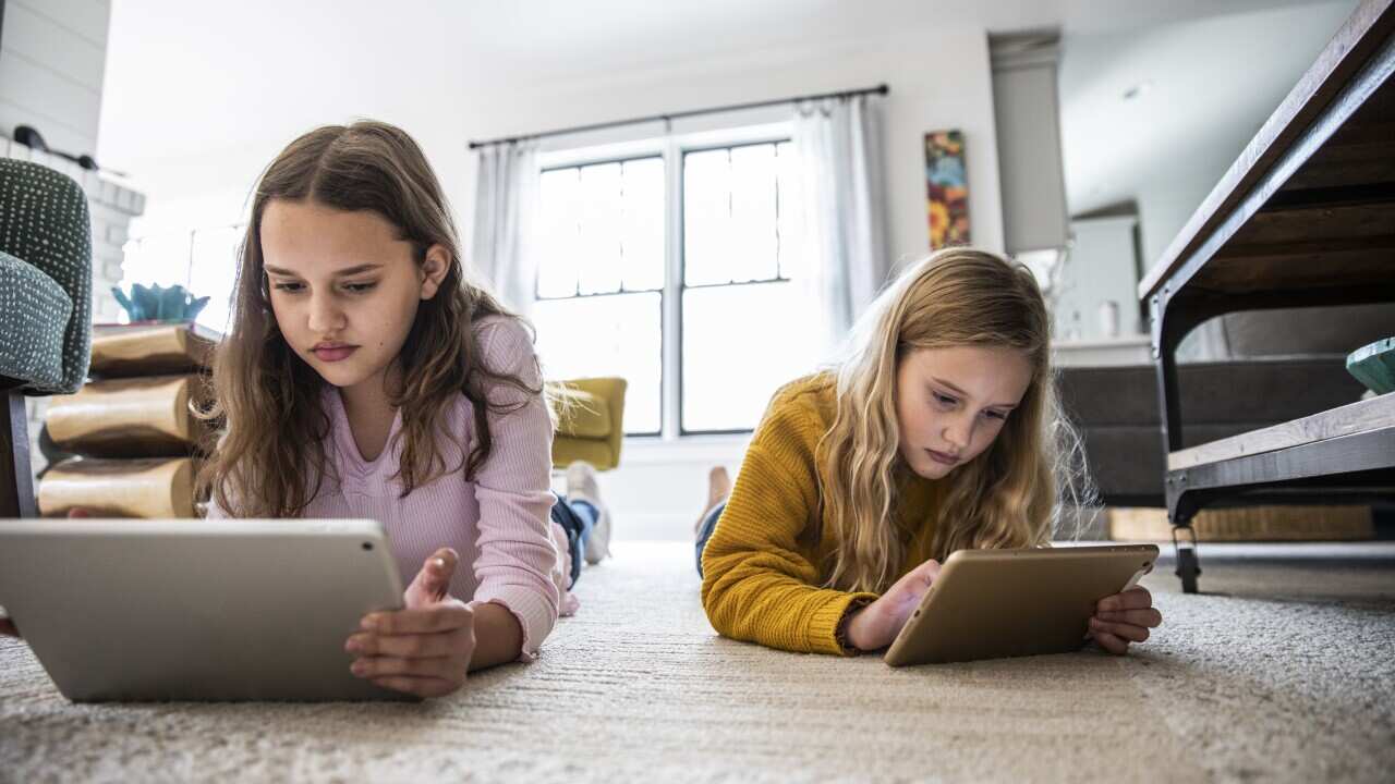 Two girls using digital tablets while lying on the living room floor.