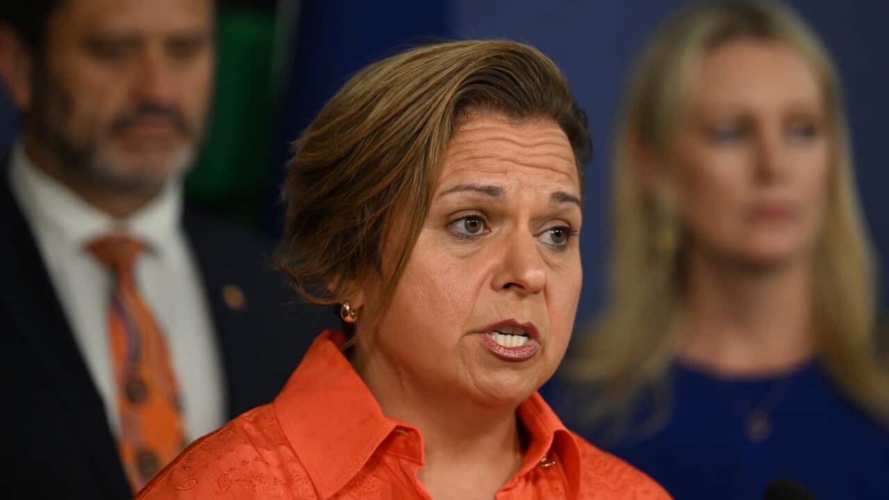 Michelle Rowland speaking while wearing a bright orange shirt, with a man and woman out of focus behind her, against a blue wall.