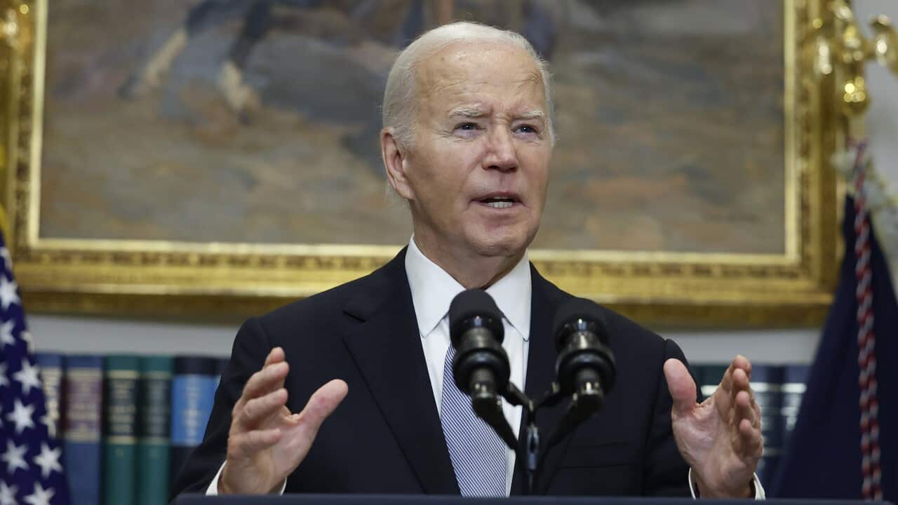 A man in a dark suit with white hair stands behind a lecturn and microphones