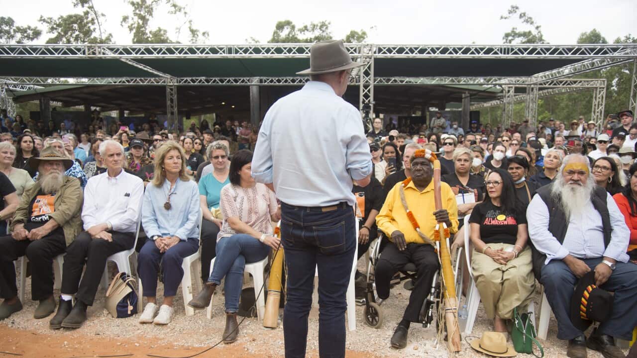 Anthony Albanese, wearing jeans, a light blue shirt and a broad-brimmed hat, and with his back to camera, talks to an audience seated outside.
