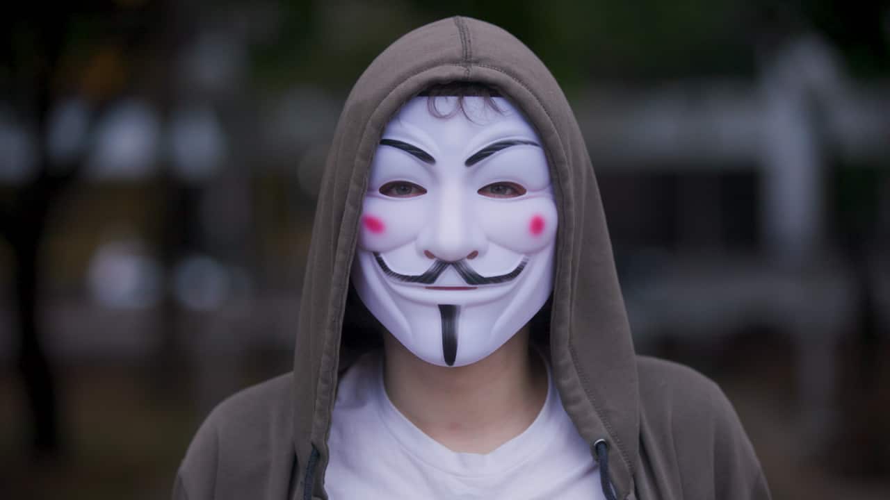 Man wearing a mask and hoodie stares directly into camera.