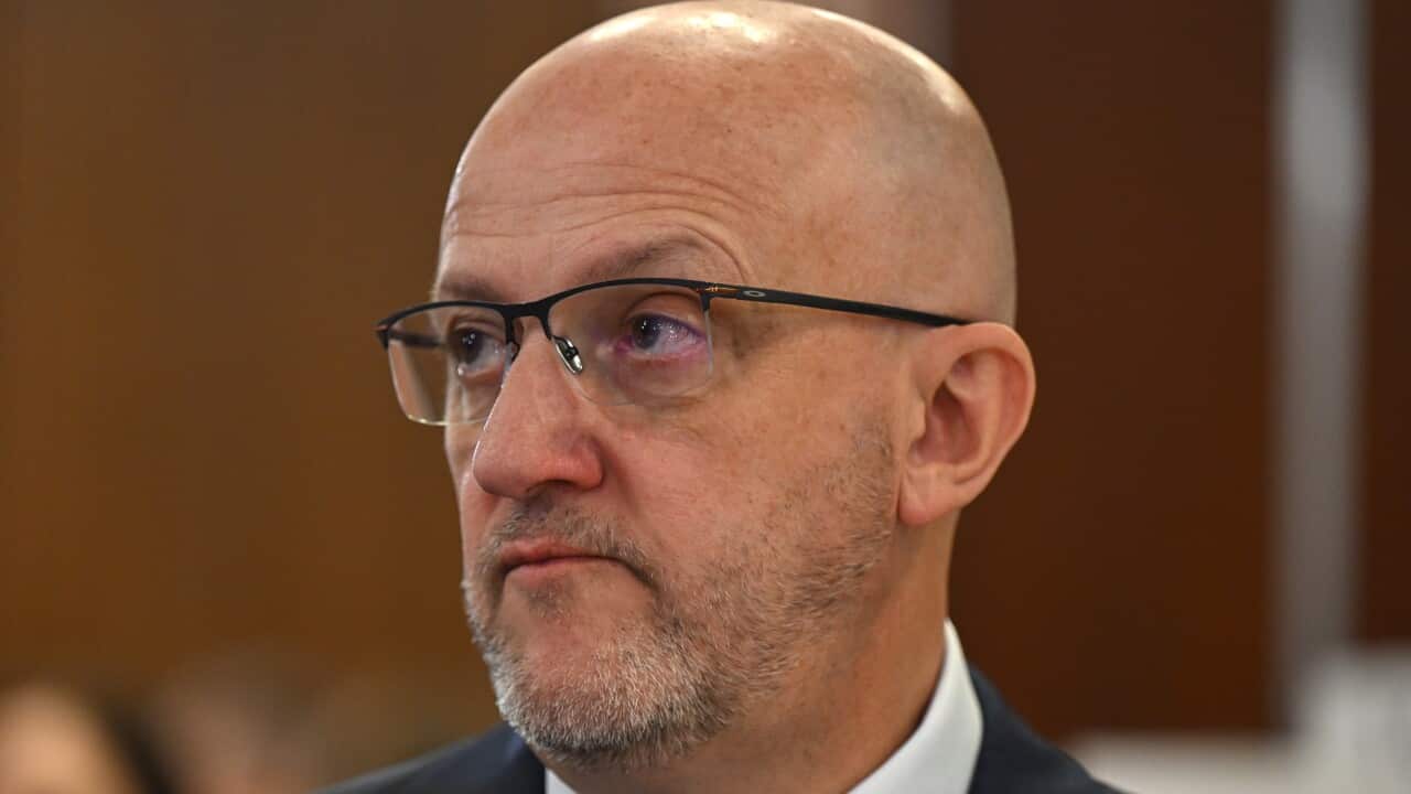 A close-up photograph of a bald, middle-aged man with a goatee and glasses, wearing a suit and tie, looking off to the side with a serious expression.