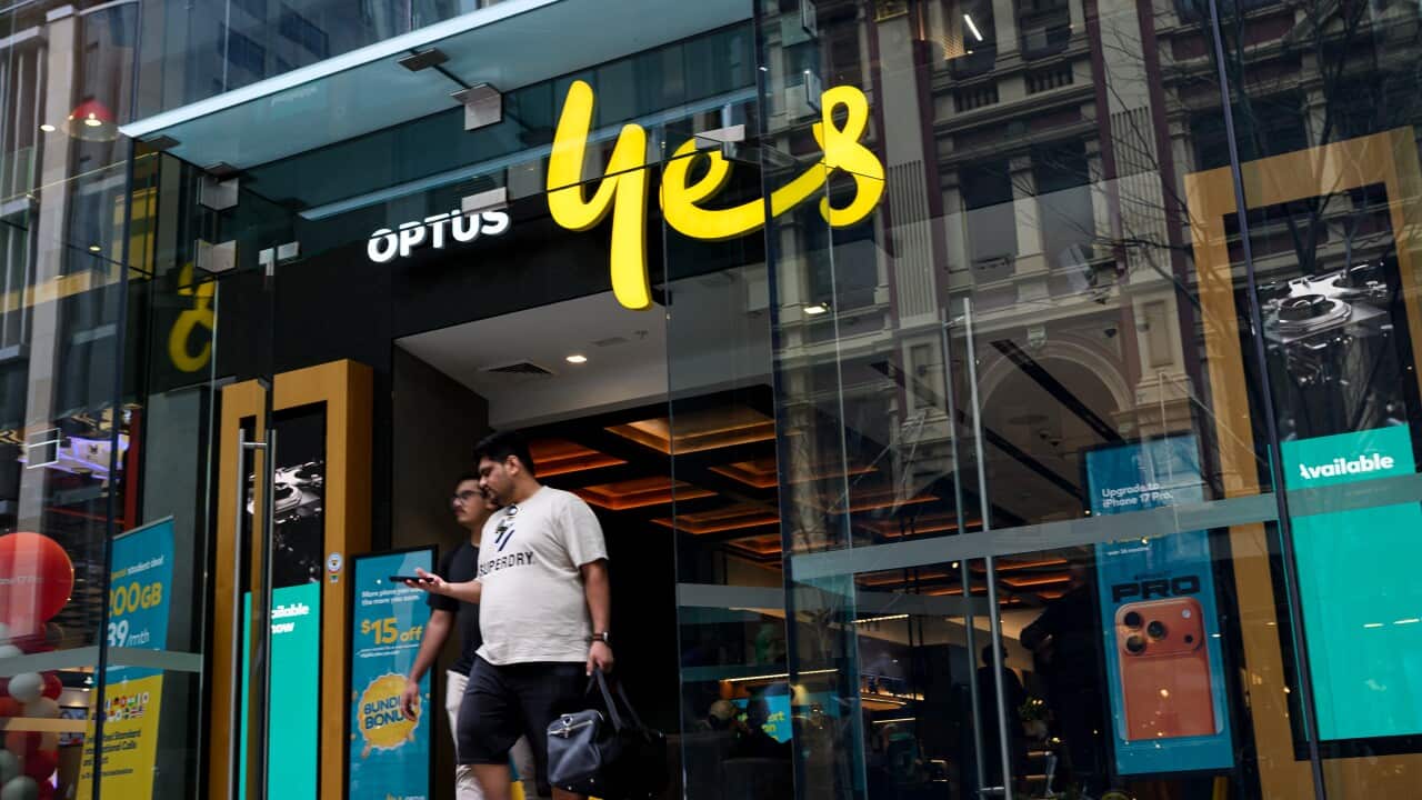 A wide-angle street-level shot shows the entrance to an Optus store with the company's “yes“ logo in bright yellow above the door, as two men walk out of the glass entrance.