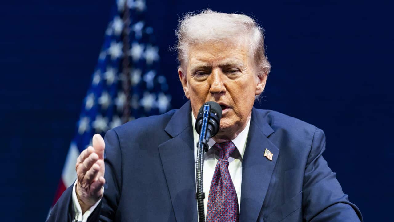Donald Trump, wearing a blue suit, speaks into a microphone in front of an American flag.