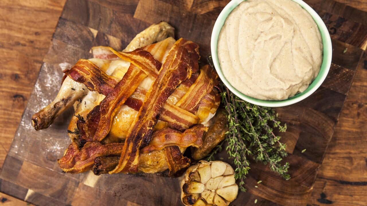 Seen from overhead, a cooked roast chicken, decorated with bacon, sits on a board. Creamy sauce sits in a small bowl alongside.