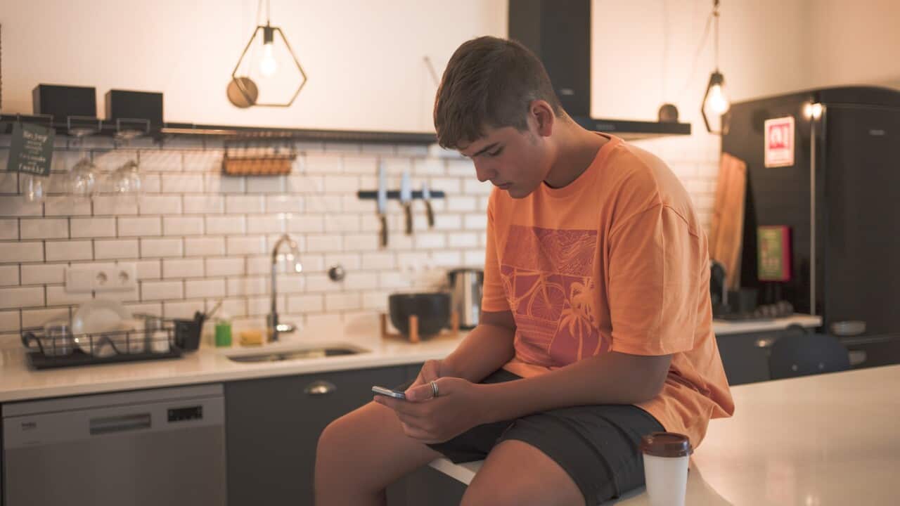 Teenage boy sitting at the kitchen table interacting with his smart phone while having a takeaway coffee