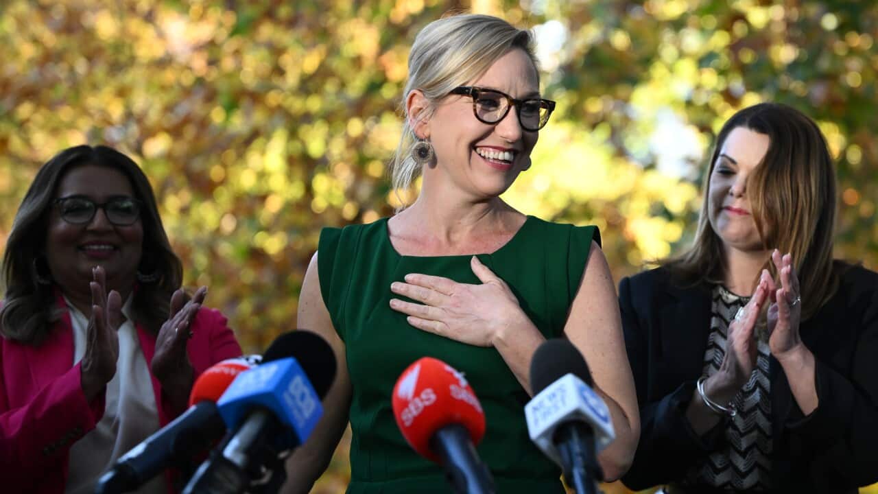A woman wearing a green dress standing in front of microphones. Two women standing behind her are clapping.